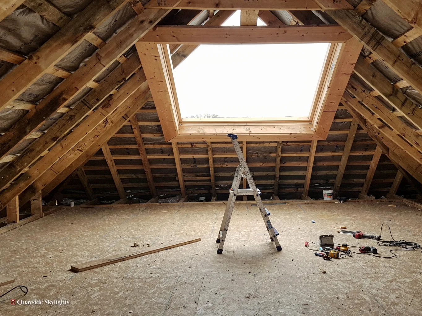 Loft under conversion with a roof window between exposed rafters, a stepladder and tools on boarded floor