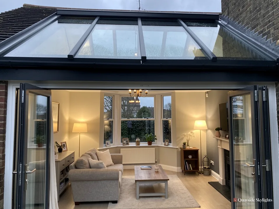 Living room with a dark-framed glazed roof and open bifold doors, seen from the garden
