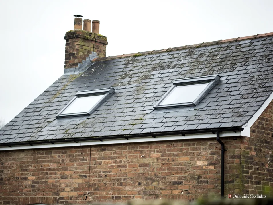 Two roof windows side by side in a slate roof below a brick chimney stack