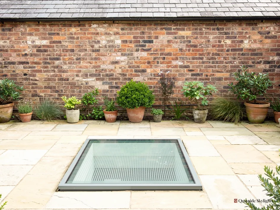 Walk-on glass panel set into a stone-paved courtyard with potted plants against a brick wall