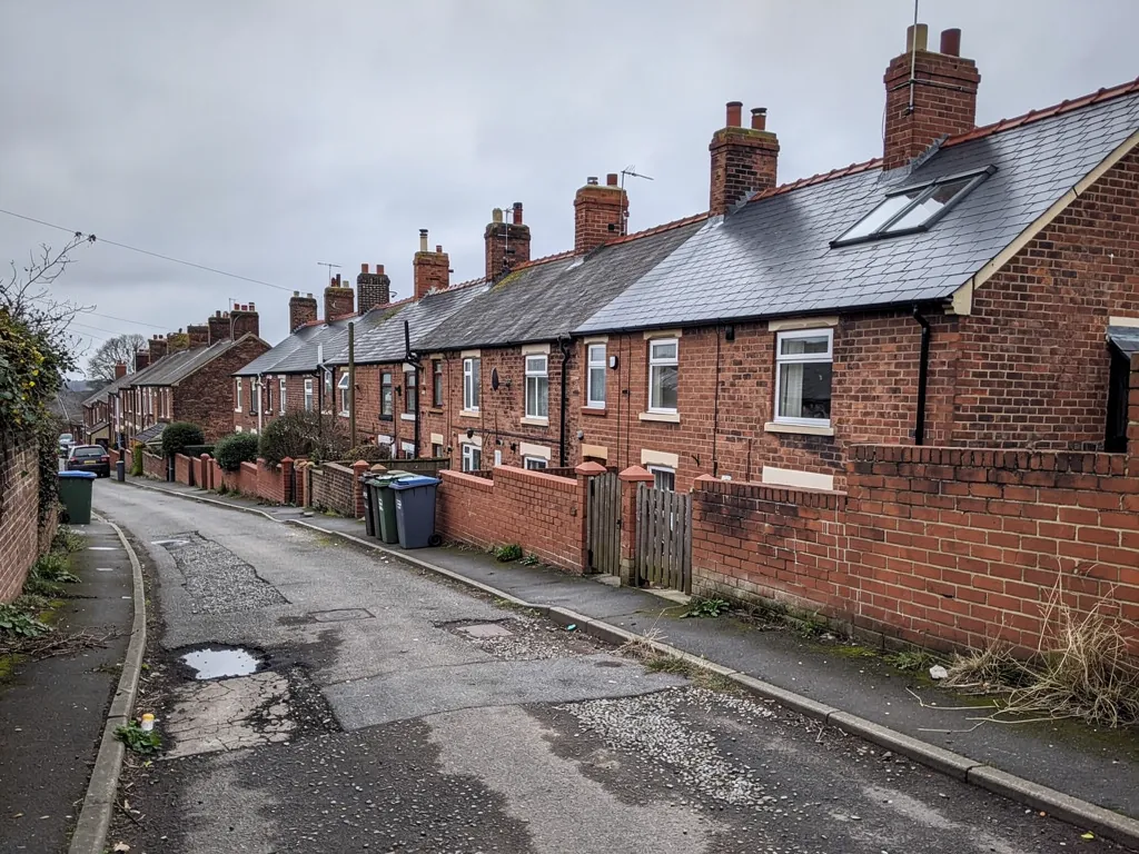 Brick terraced street with potholes and wheelie bins on a grey day