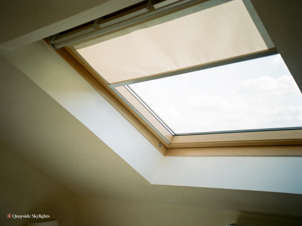 Roof window with a pale fabric blind half drawn, seen from below in a sloping ceiling